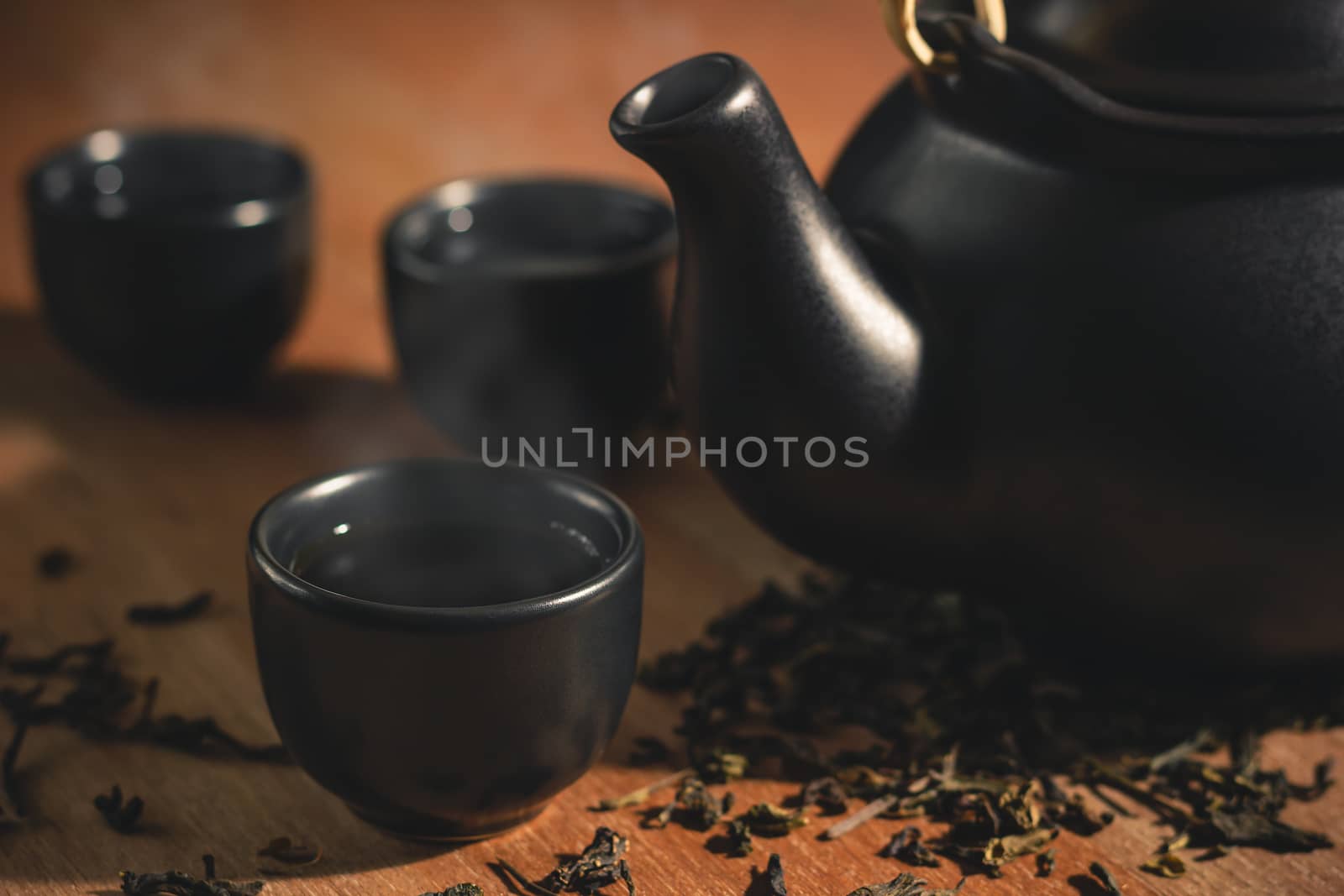 Glass of tea with hot smoke is placed beside a ceramic teapot and dried tea leaves scattered on the wood background.