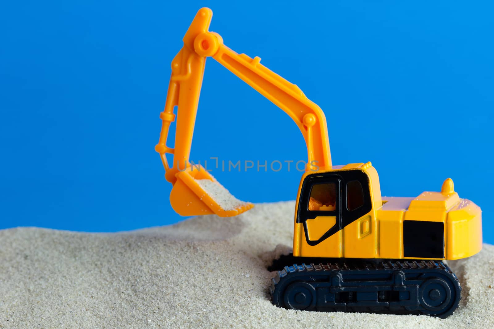 Yellow toy backhoe is digging sand on the blue background. Conce by SaitanSainam