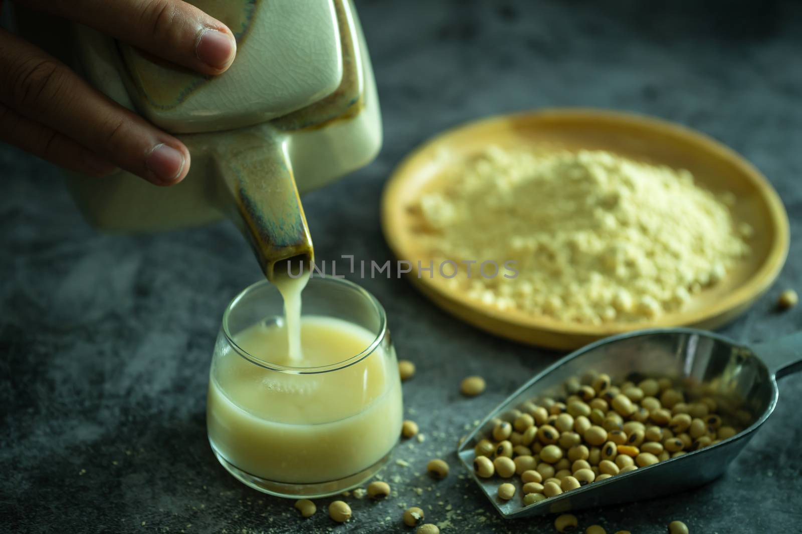 Hand holding the kettle to pour soy milk into the glass. Soybean powder is crushed with soybean seeds scattered on the table and morning light.