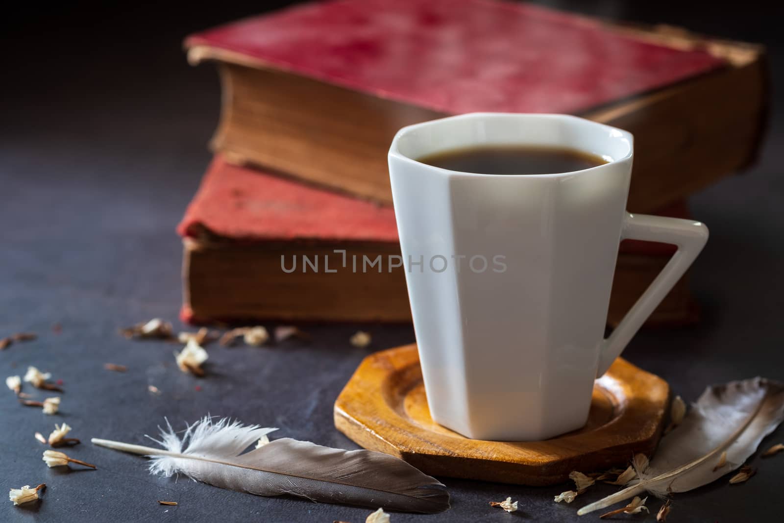 Black coffee in white cup and old books with feather and dried flower petals placed on the marble table and morning sunlight.