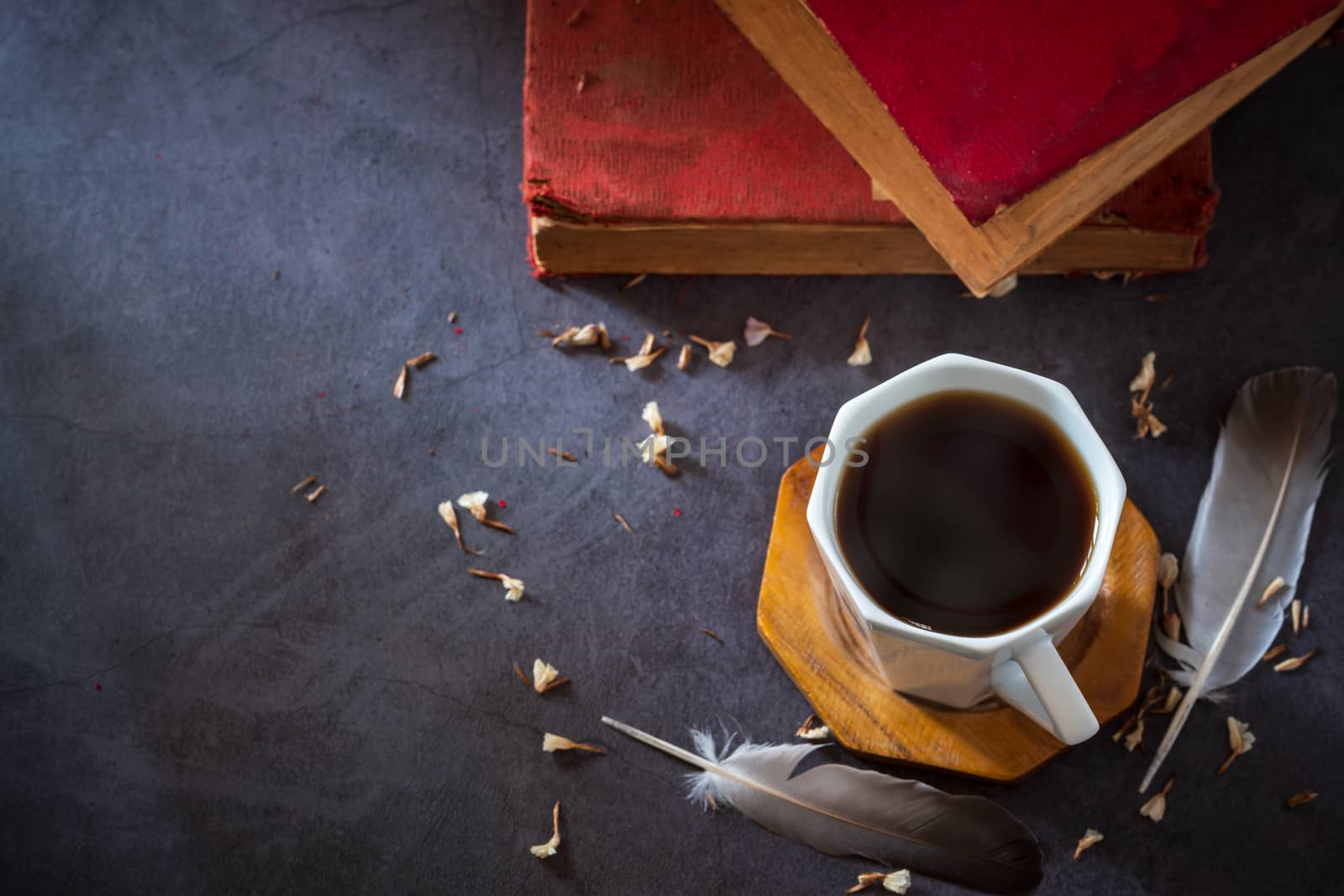 Black coffee in white cup and old books with feather and dried flower petals placed on the marble table and morning sunlight.