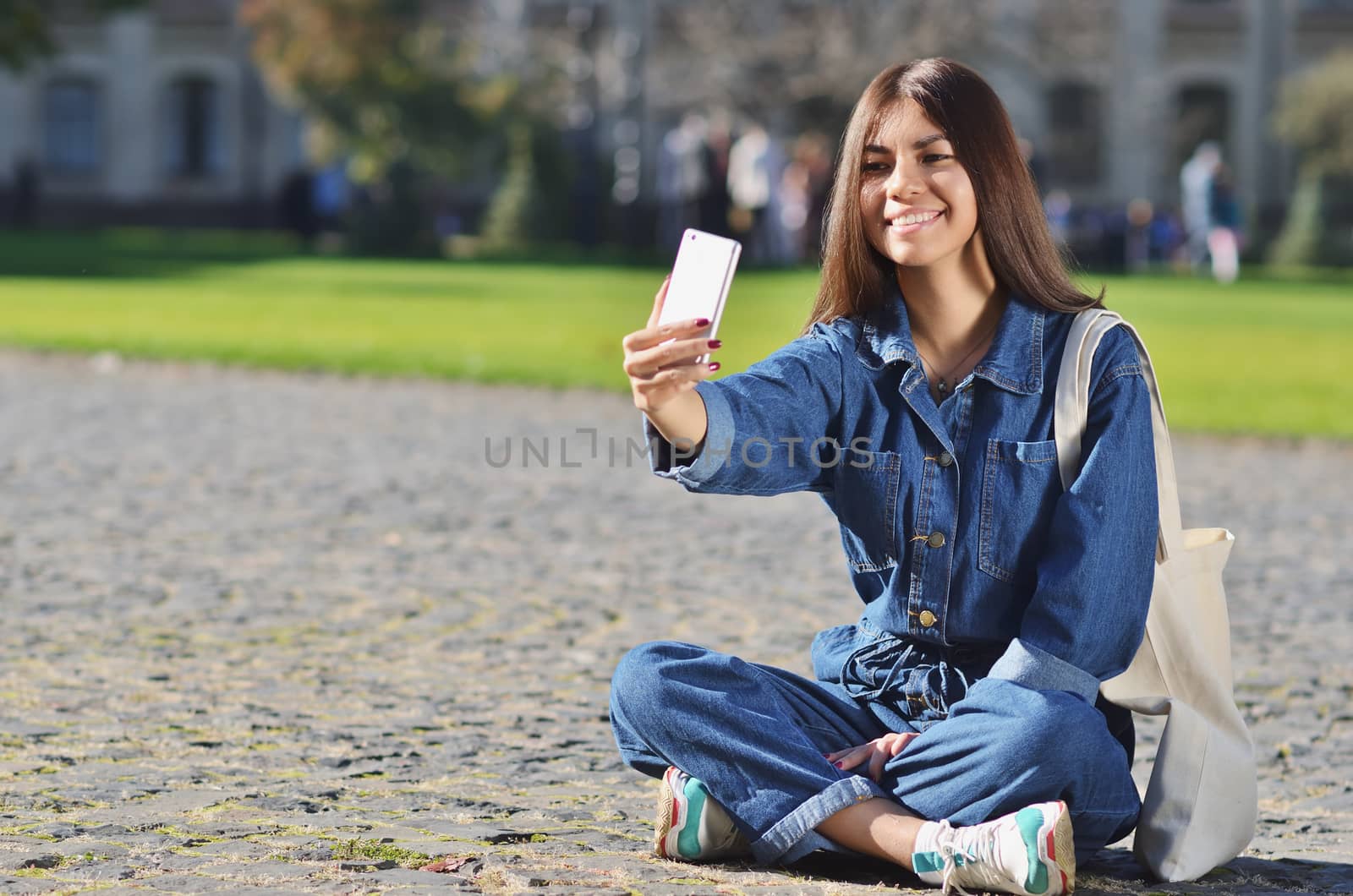 Cheerful young woman is sitting in the campus and taking a selfie, dressed in a denim jacket and sneakers