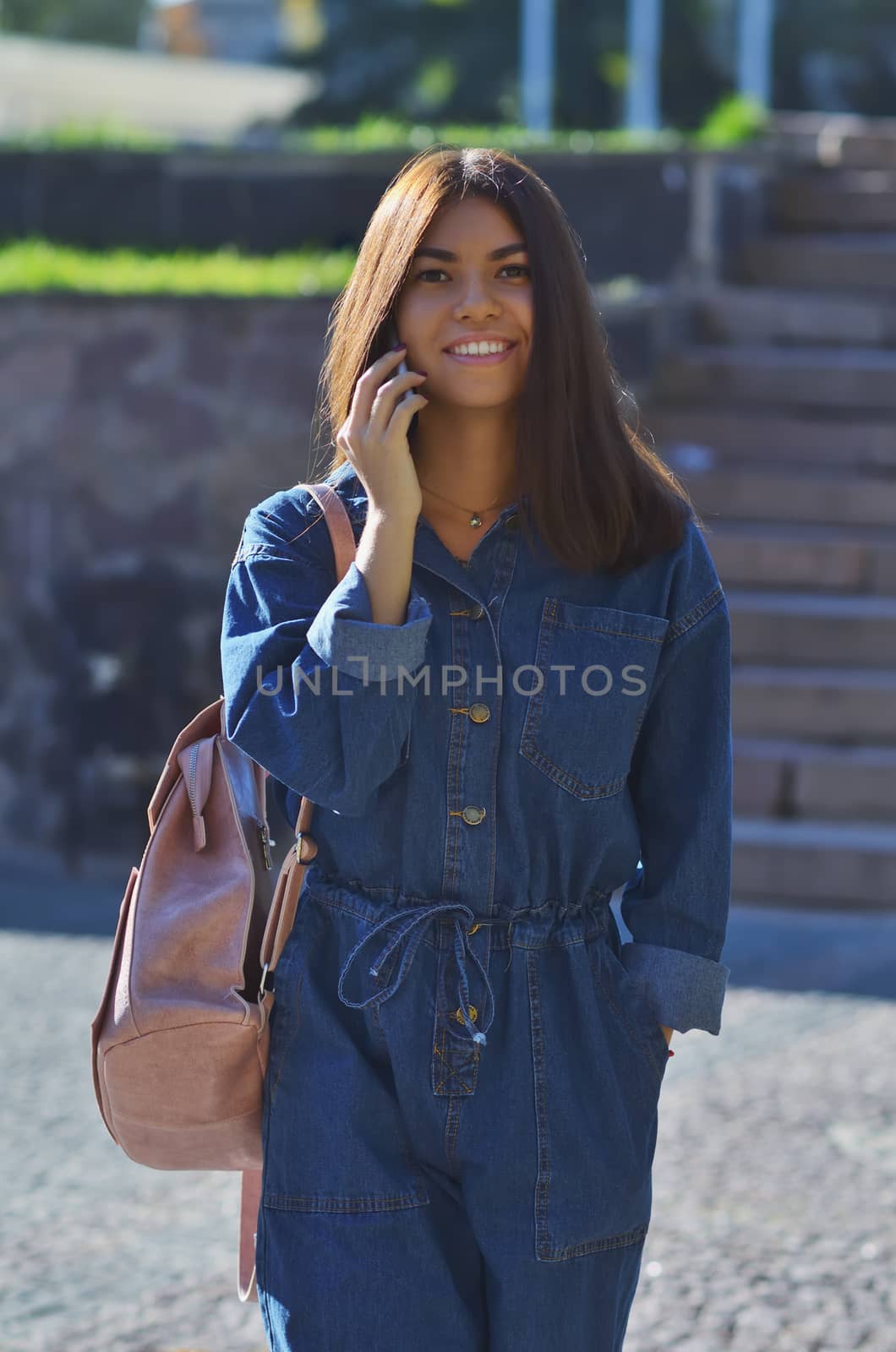 A young girl smiles, walks around the city with a pink backpack talking on the phone, dressed in a denim suit and light sneakers