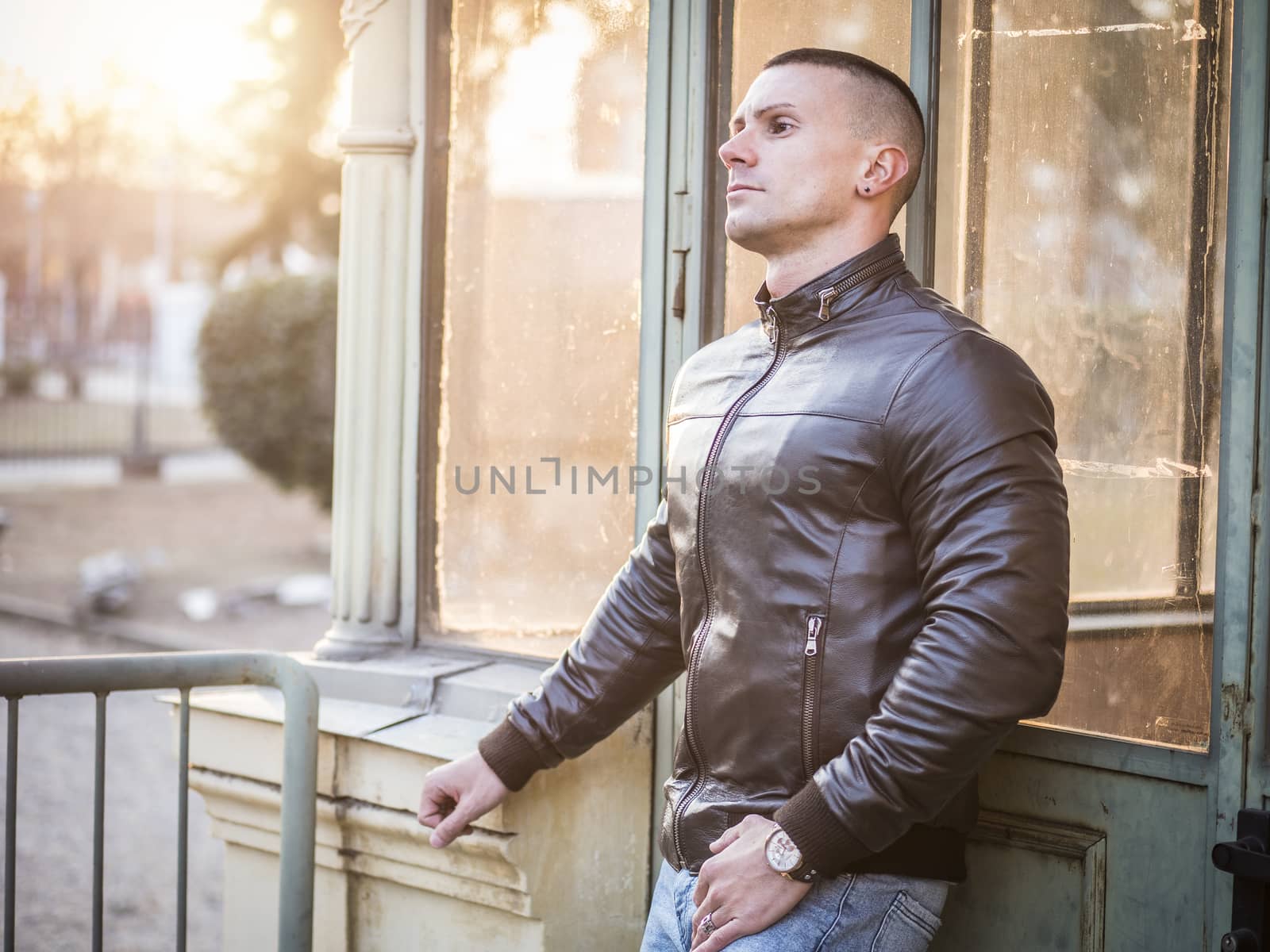 One handsome young man in urban setting in European city, standing, wearing black leather jacket and jeans at sunset