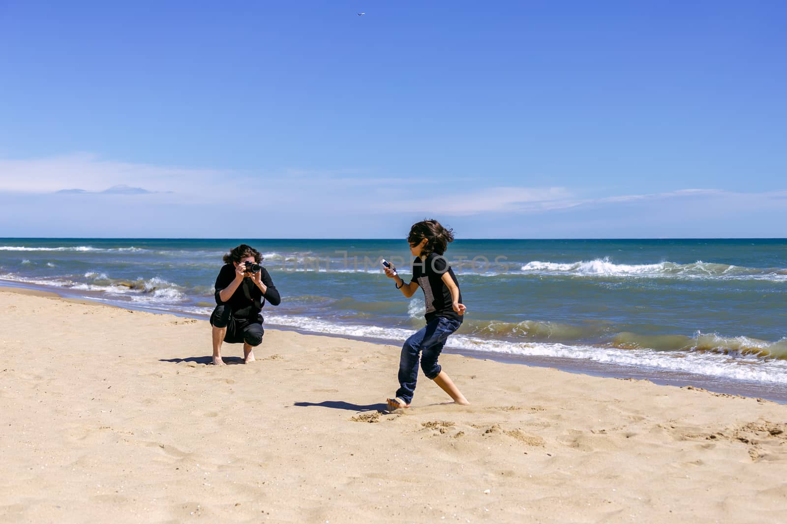 Professional photographer with a camera shoots a running teenager on a sandy beach of the ocean coast on a sunny spring day