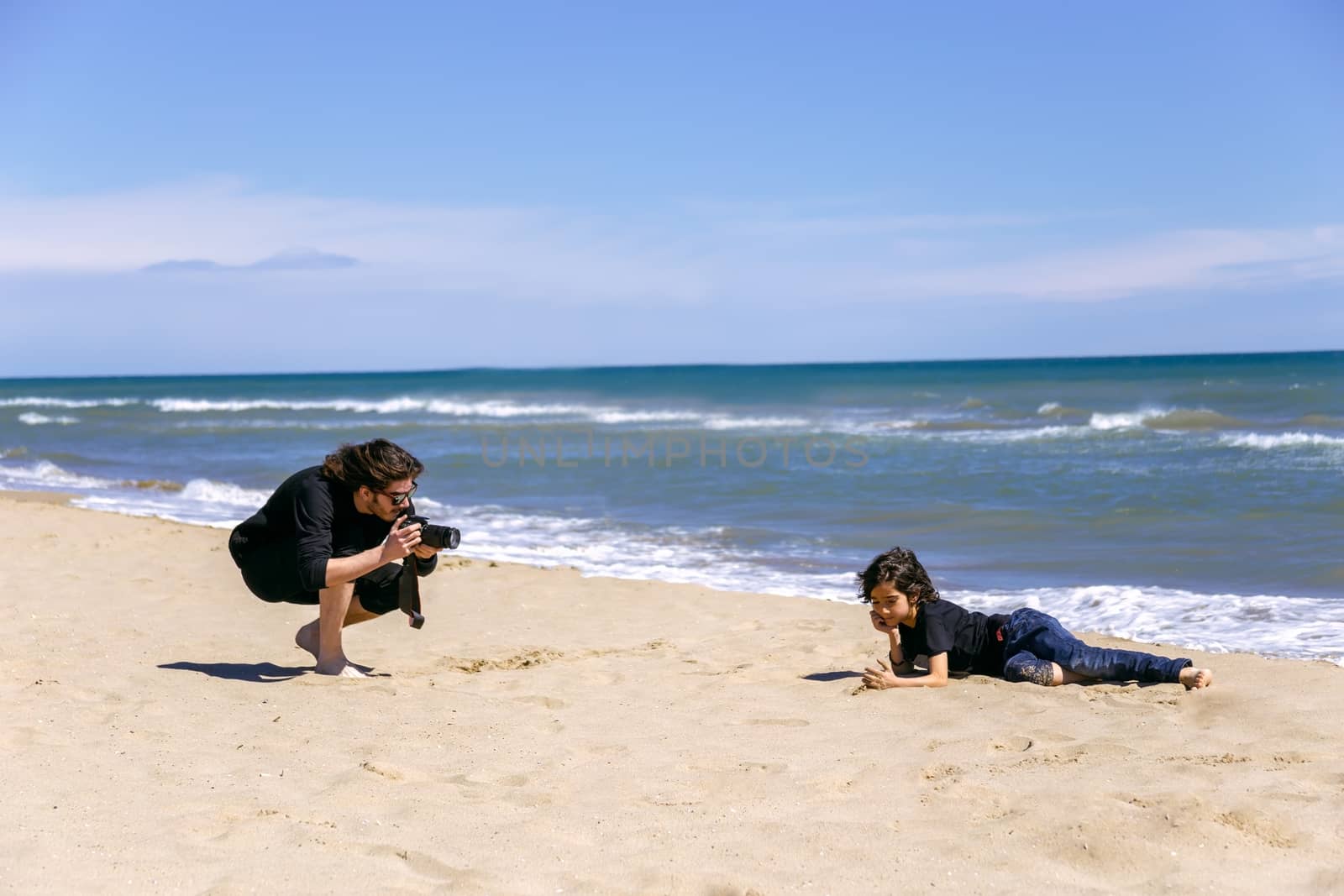 Professional photographer with photo camera shooting teenage boy lying on sandy beach of ocean coast