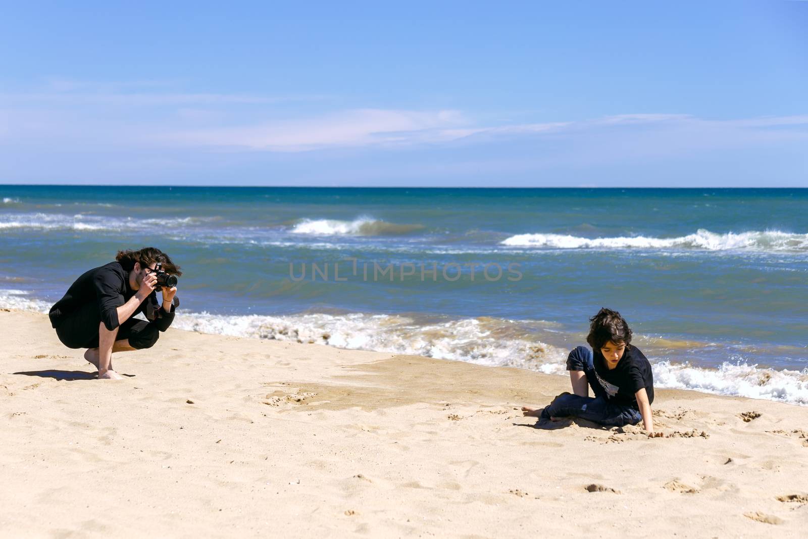 Professional photographer with photo camera shooting teenage boy lying on sandy beach of ocean coast