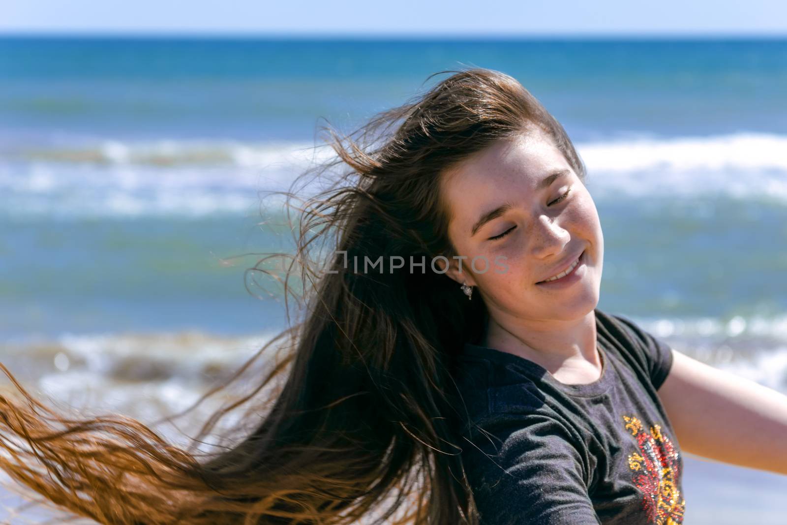 Beauty Sunshine Girl Portrait. A teenager girl with long brown hair flying in the wind smiles happily, closing her eyes and exposing the sun to her face against the backdrop of the sea.