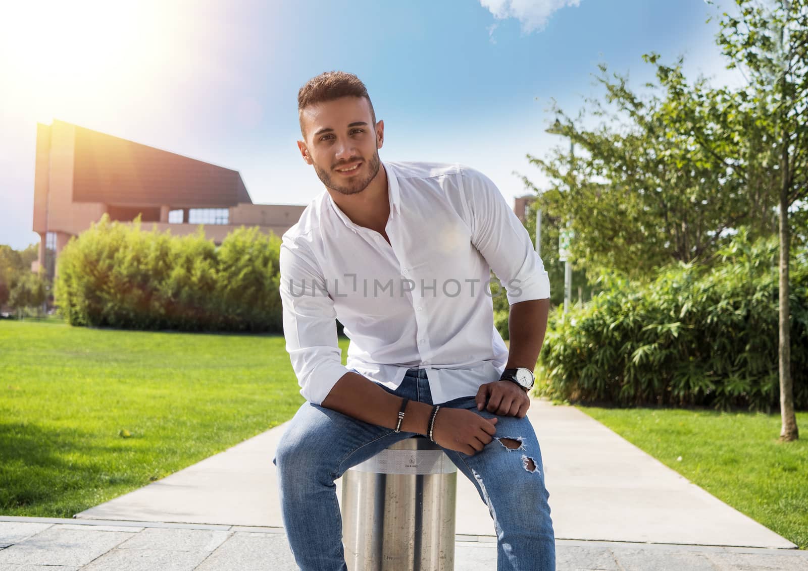 Attractive handsome young man in city park, sitting, in a summer day, wearing jeans and white shirt