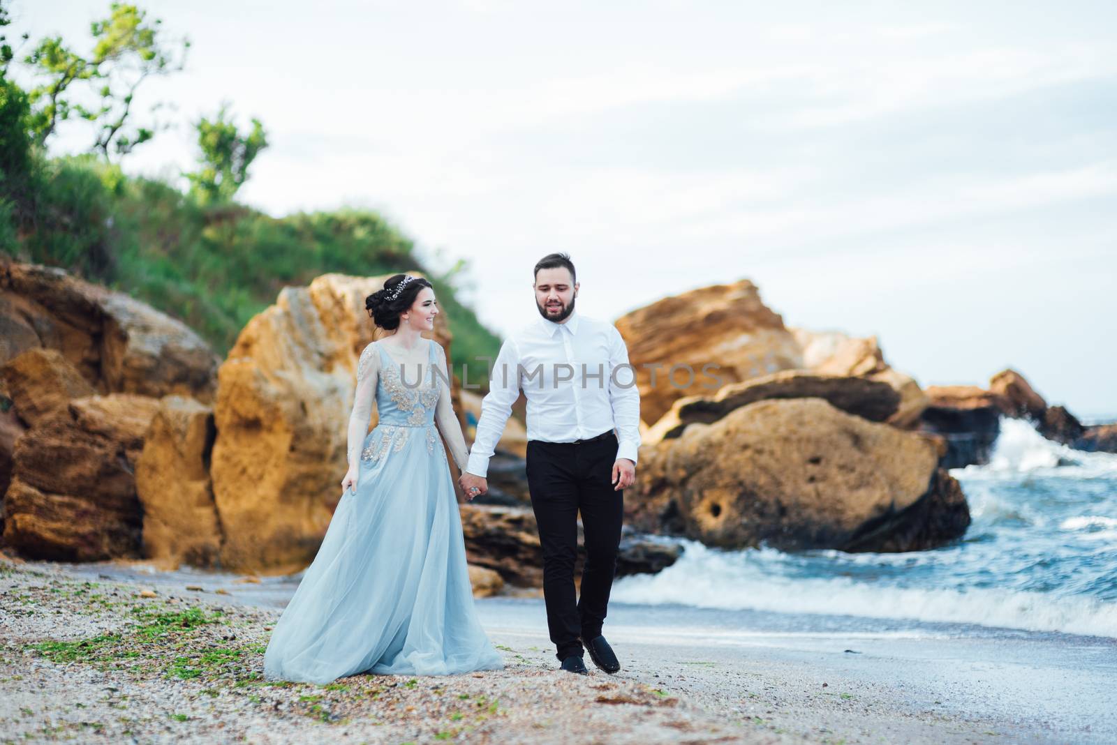 same couple with a bride in a blue dress walk along the ocean shore