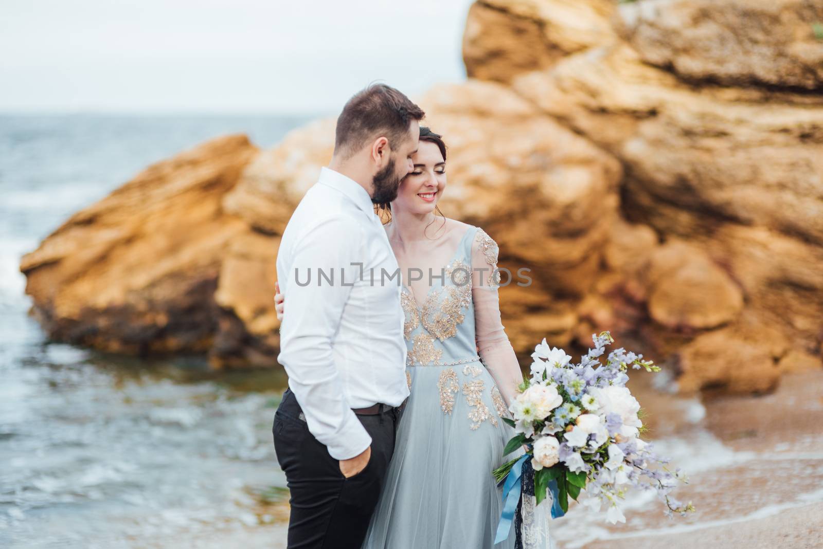 same couple with a bride in a blue dress walk along the ocean shore