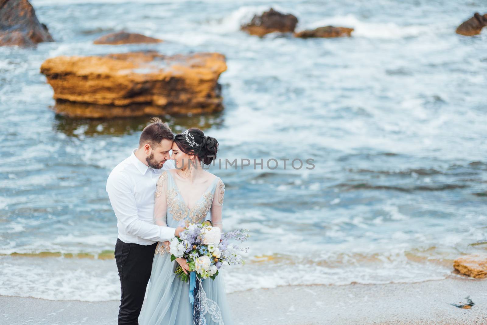 same couple with a bride in a blue dress walk along the ocean shore