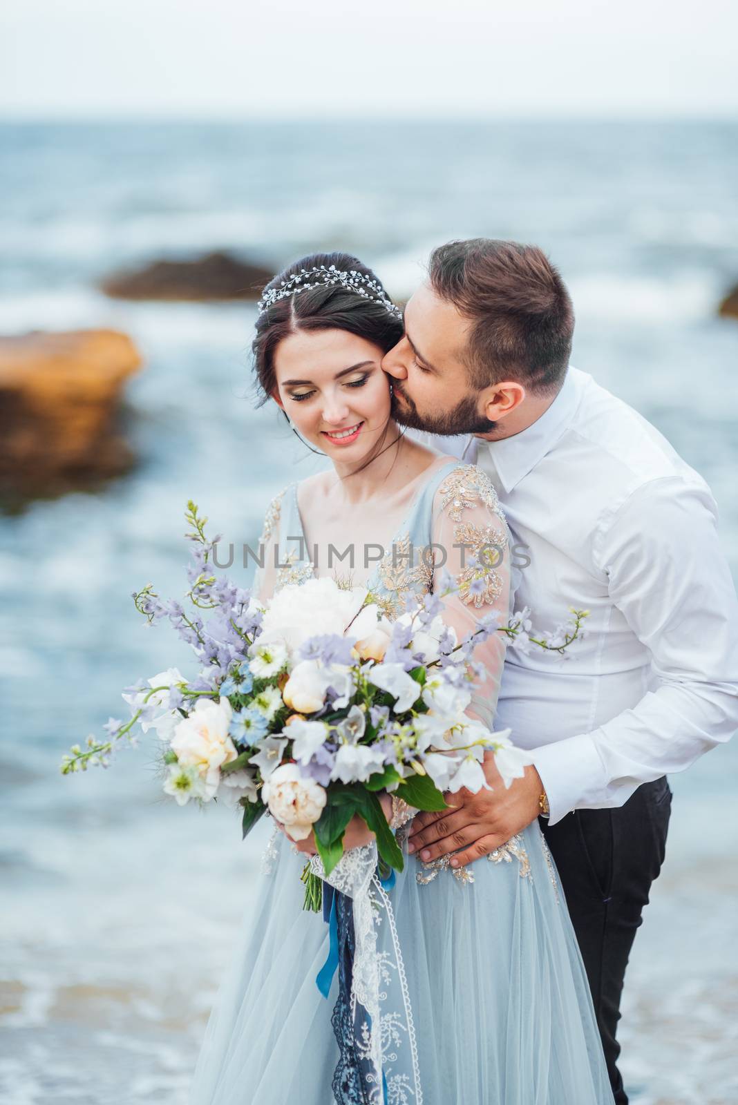 same couple with a bride in a blue dress walk along the ocean shore