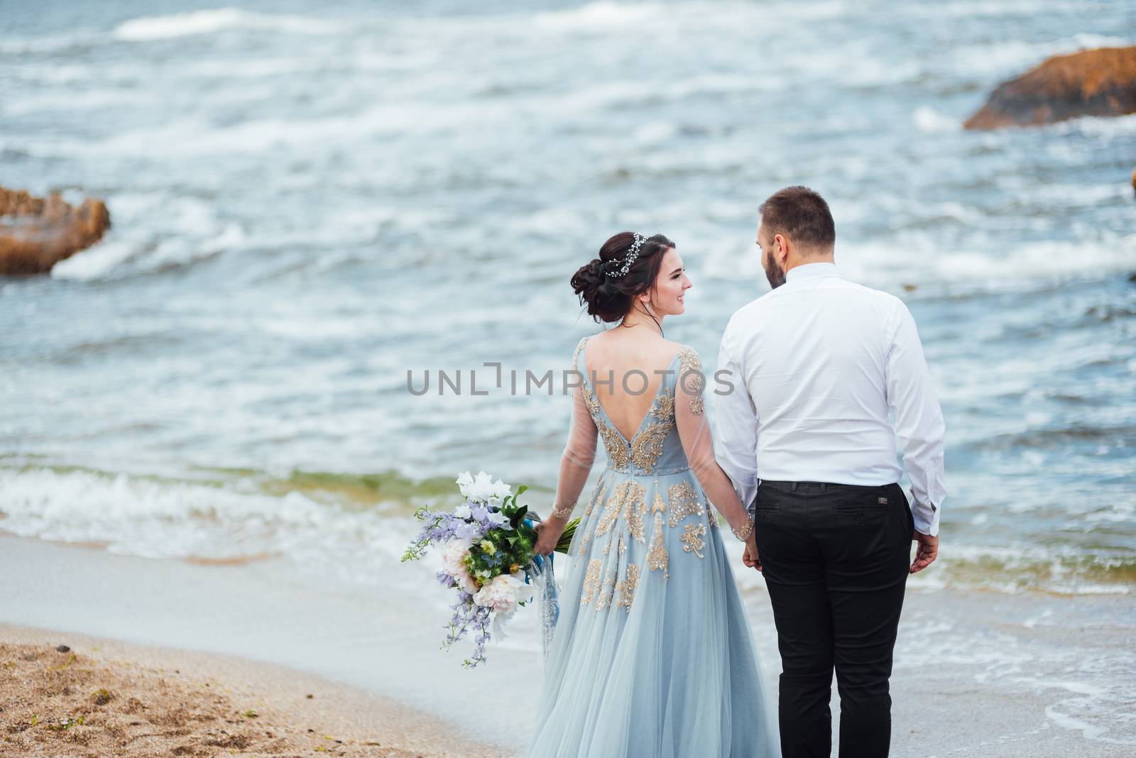 same couple with a bride in a blue dress walk along the ocean shore