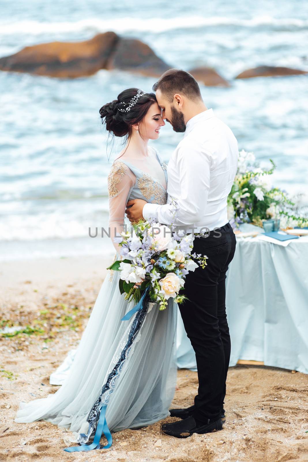 same couple with a bride in a blue dress walk along the ocean shore