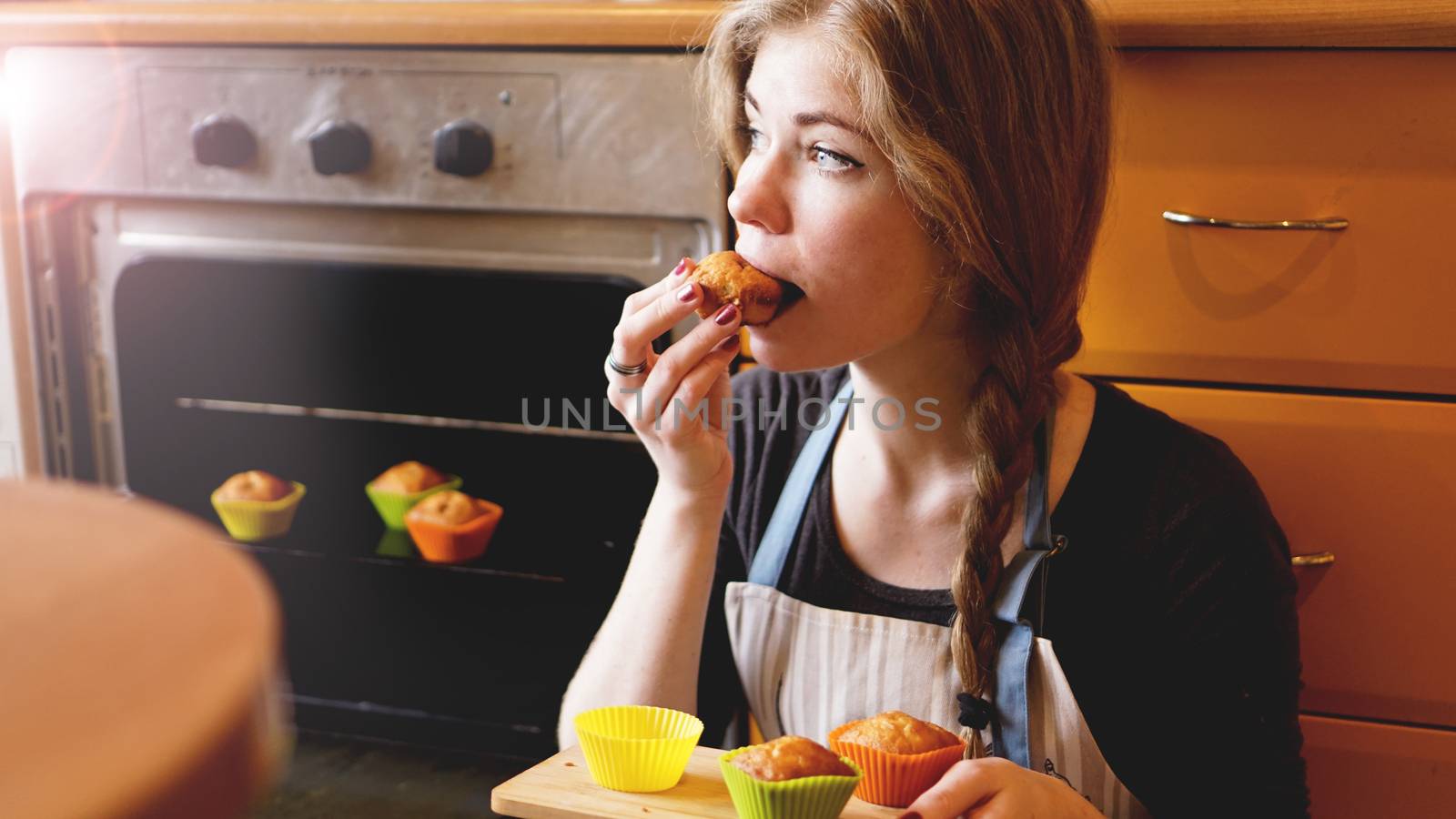 Beautiful blonde woman showing muffins while eating one in a kitchen. Cooking and home concept