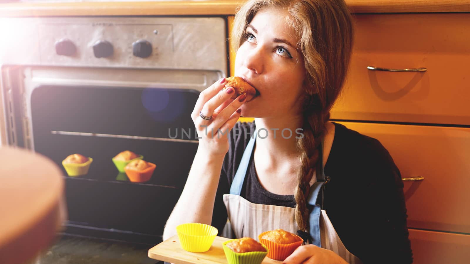 Beautiful blonde woman showing muffins while eating one in a kitchen by natali_brill