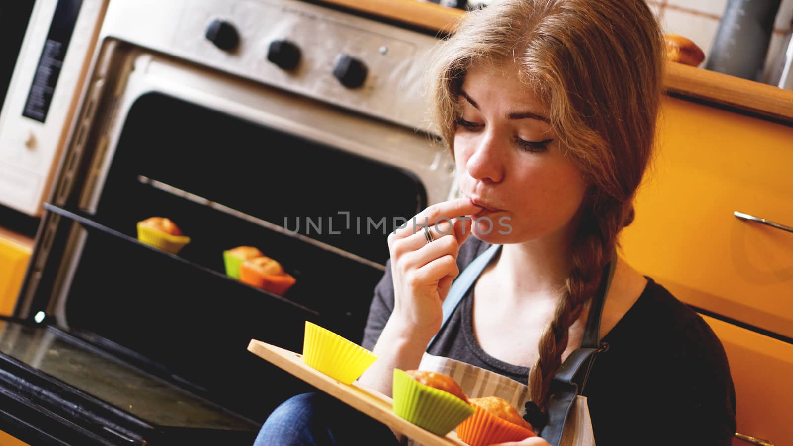 Beautiful blonde woman showing muffins while eating one in a kitchen. Cooking and home concept