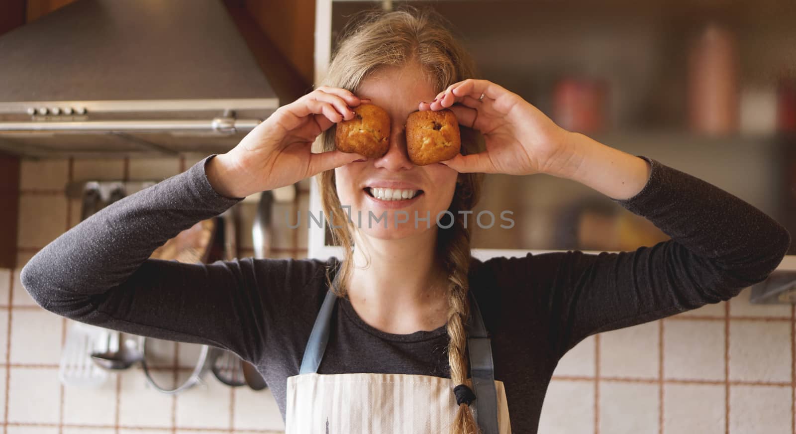 Portrait of beautiful woman in apron covering her eyes with muffins and smiling while standing in the kitchen
