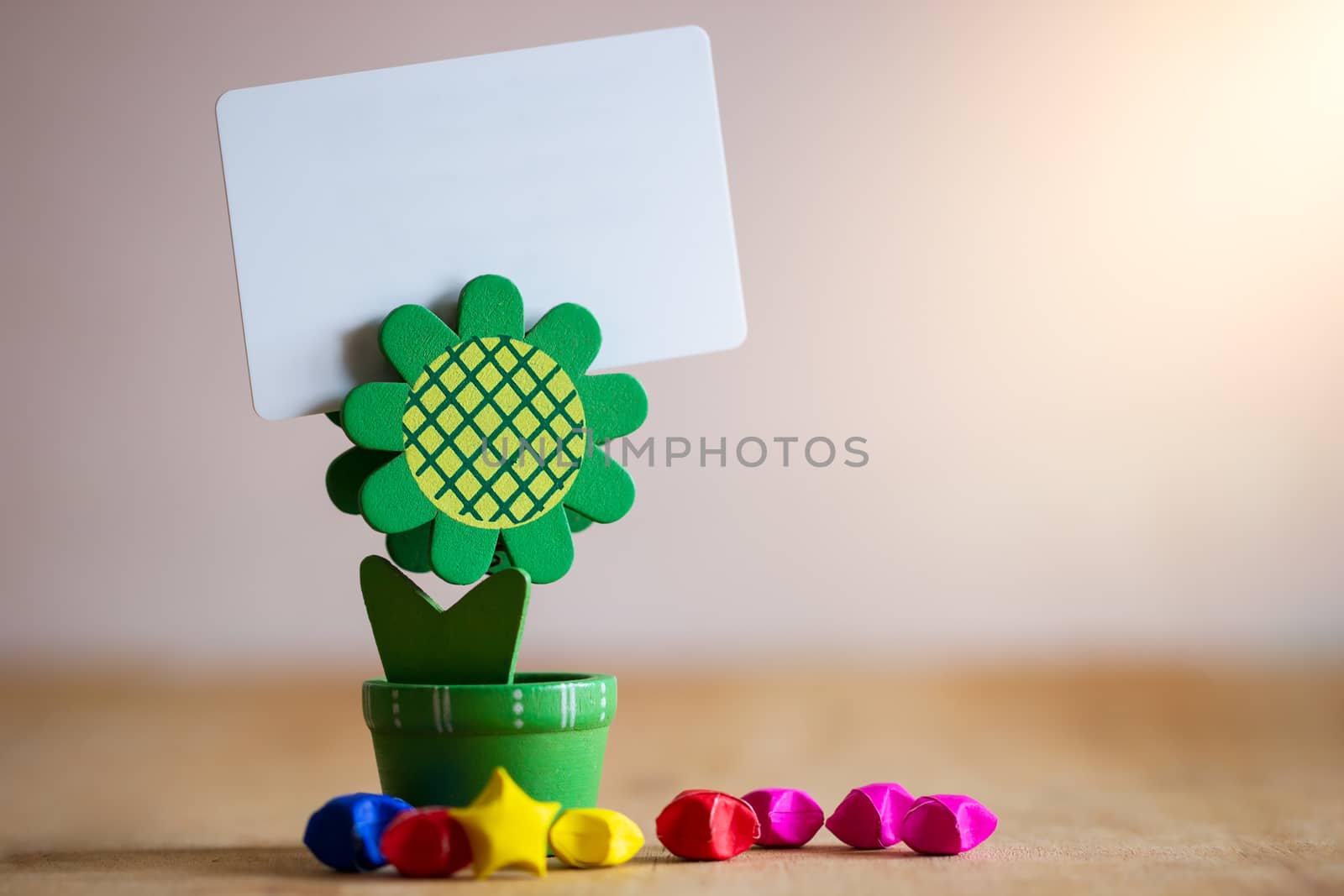 Clip holder card stand green sunflower shaped and multicolored paper stars on wooden table. White plastic blank card in morning sunlight.