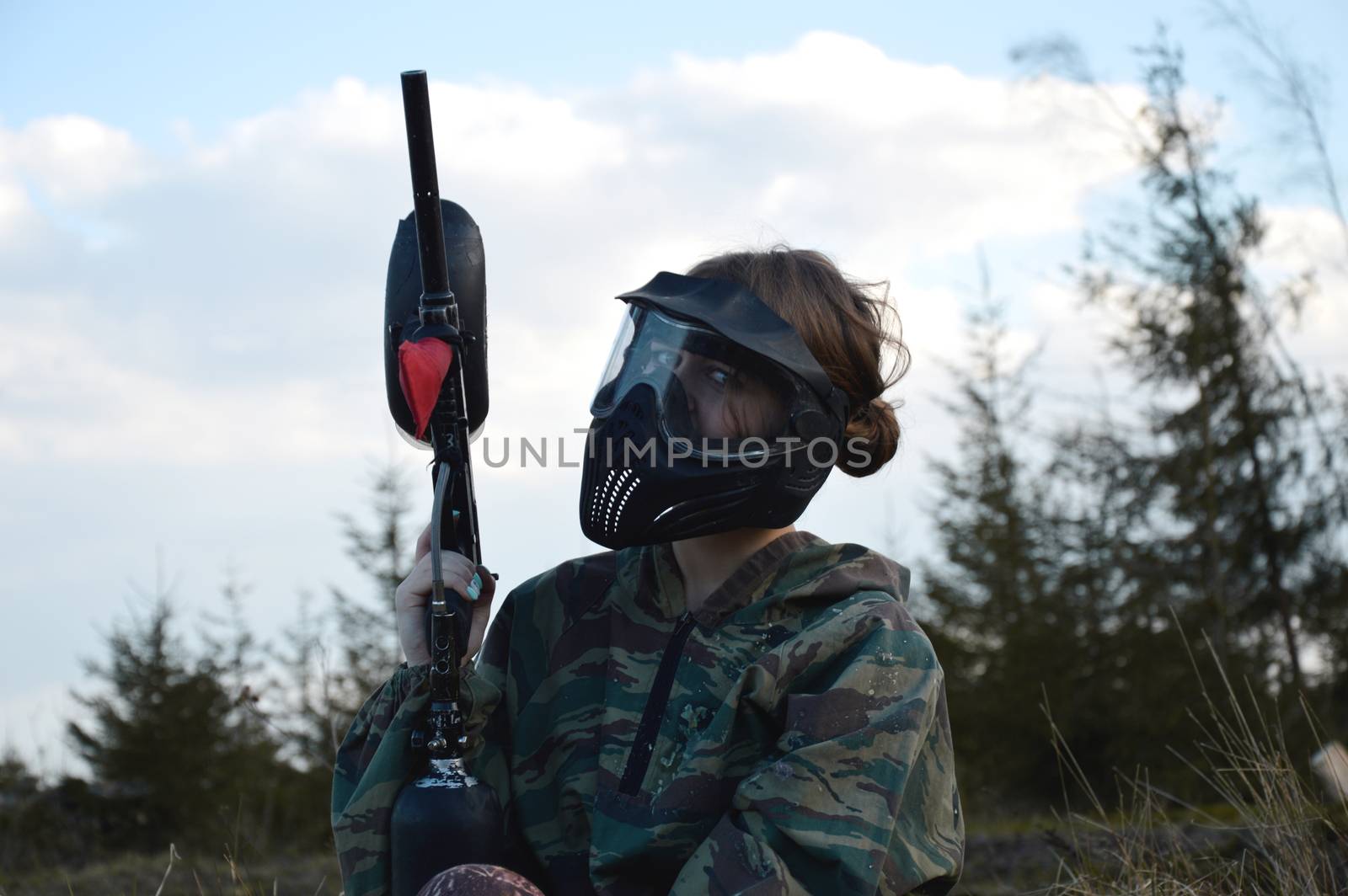 Paintball sport player girl in protective camouflage uniform and mask with marker gun outdoors