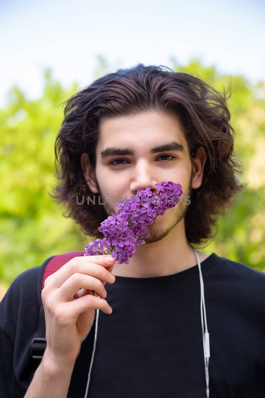 Young brunette guy smelling fresh flowers on lilac branch and looking at camera on blurred background