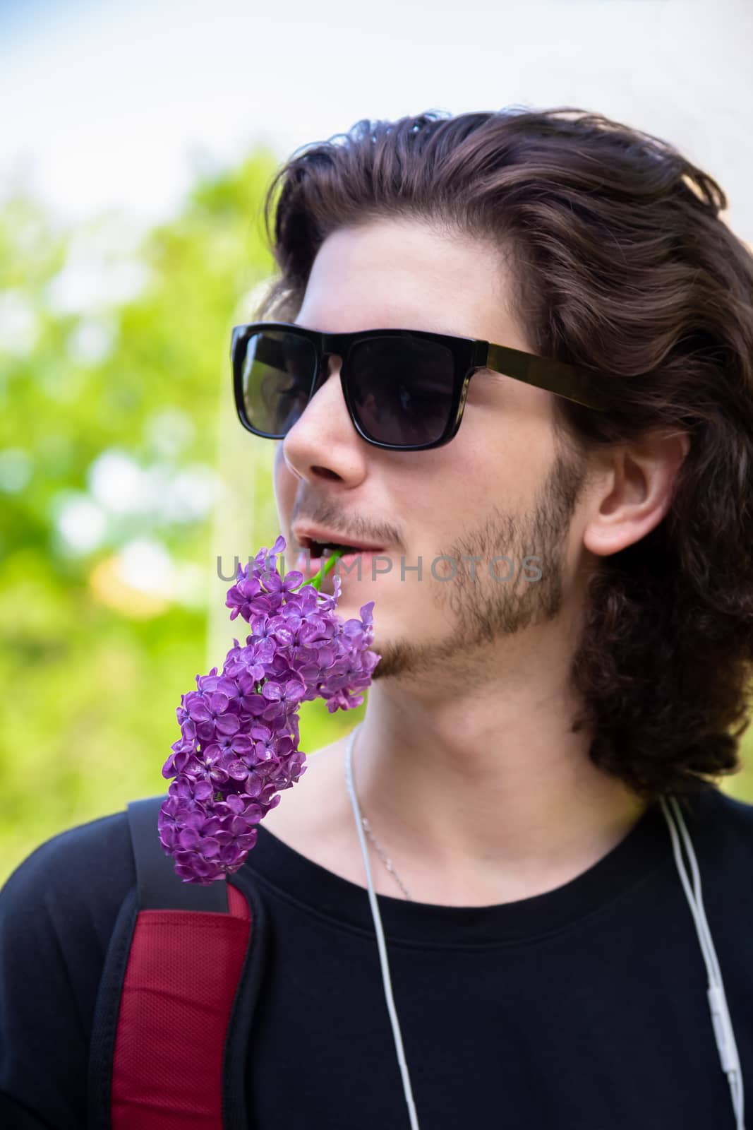 Young brunette man with long curly hair and sunglasses holds fresh flowers in his mouth on a lilac branch and looks away on a blurred background.