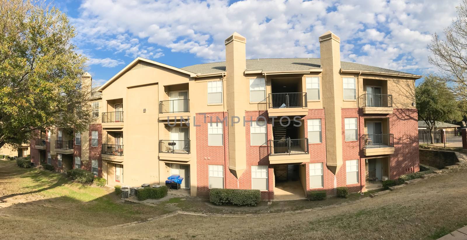 Panorama view apartment building complex with steep backyard and oak trees in suburban Dallas, Texas, USA. Low angle view of multi-stories rental real estate with covered parking at sunset cloud sky