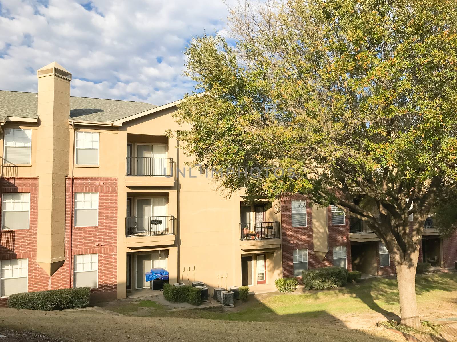 Apartment building complex with steep backyard and oak trees in suburban Dallas, Texas, USA. Low angle view of multi-stories rental real estate with covered parking at sunset cloud sky