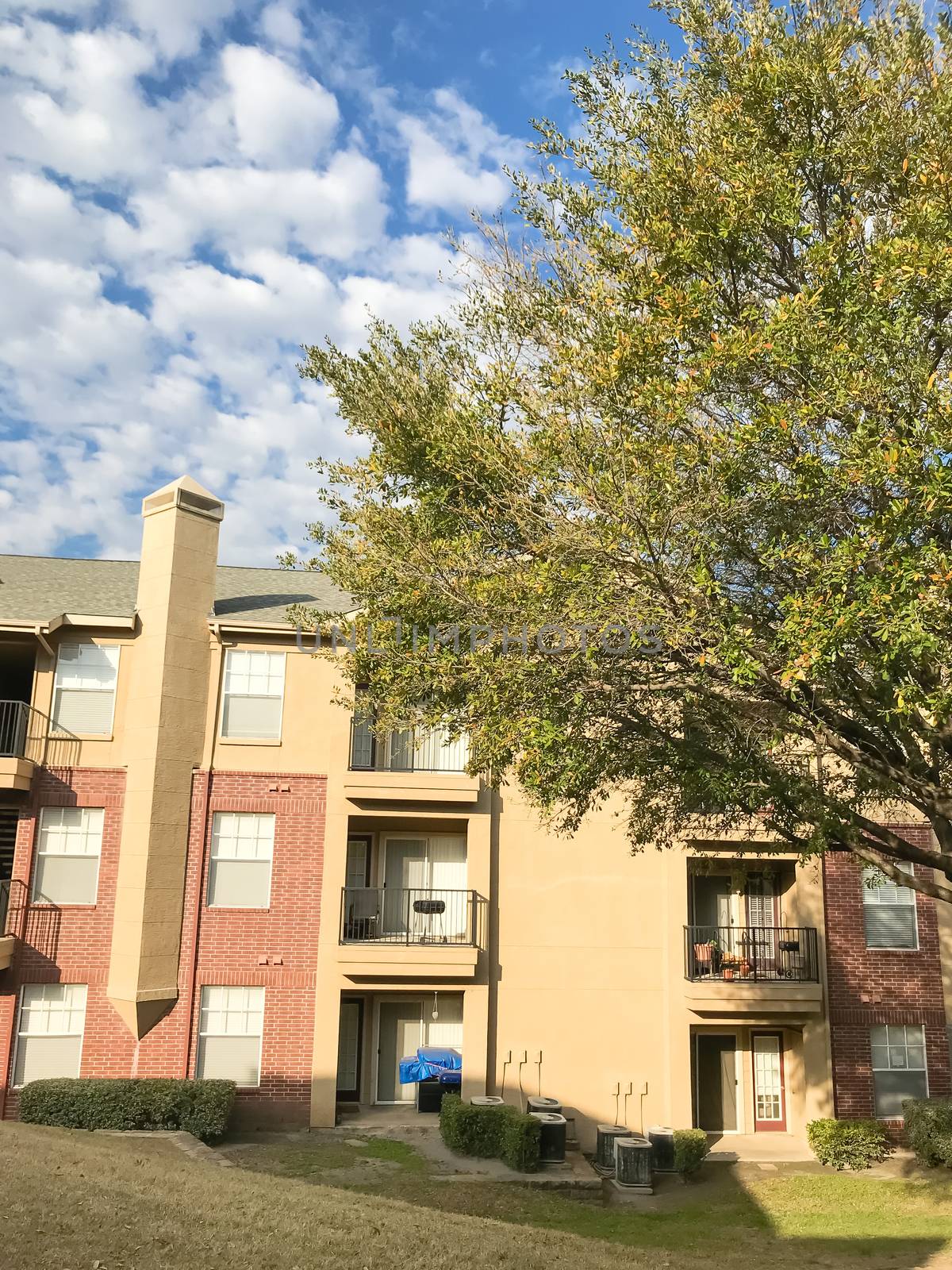 Apartment building complex with steep backyard and oak trees in suburban Dallas, Texas, USA. Low angle view of multi-stories rental real estate with covered parking at sunset cloud sky