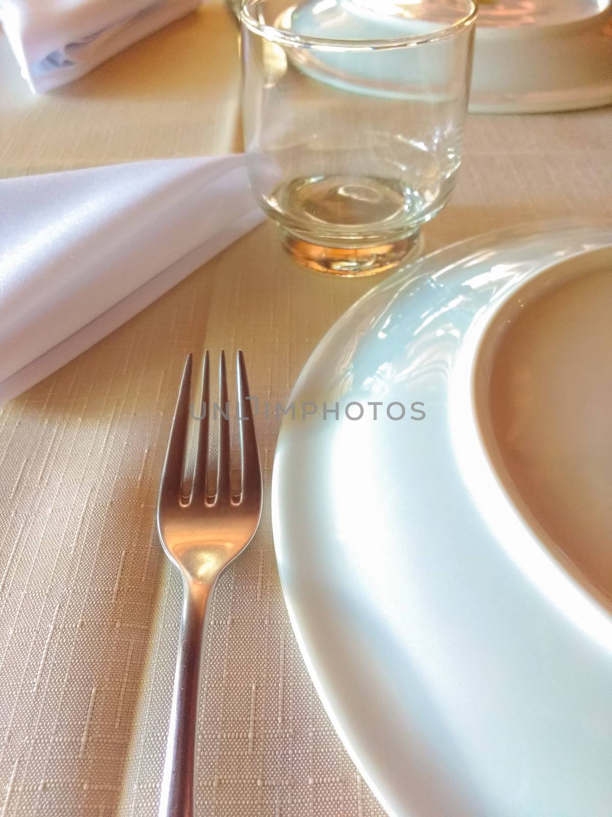 Linen tablecloth, ready to serve dinner with fork, glass, plate and napkin, under soft light in restaurant