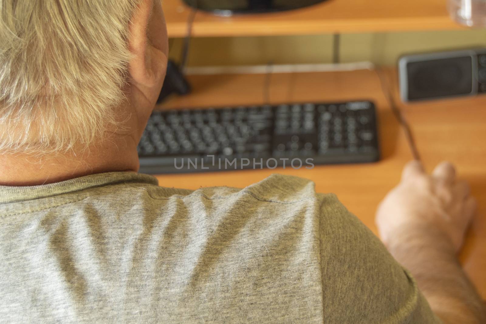 An elderly gray-haired man uses a computer mouse, work at home for the disabled, training pensioners to work on a PC, a view from the back.