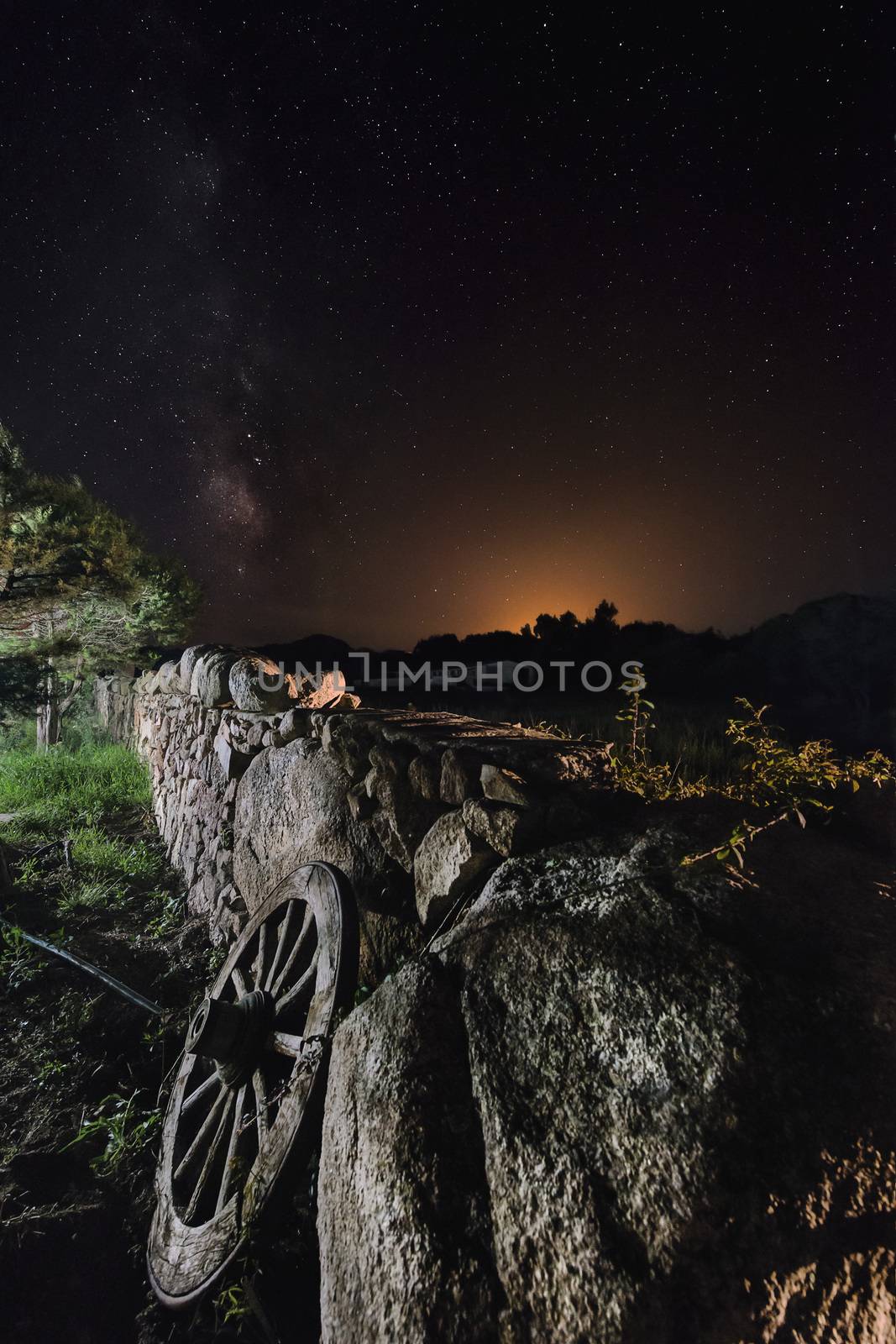 A wagon wheel is supported on the long wall of large stones of a farm. The dark night sky is full of stars and behind the mountains you can see the reflection of the lights of a nearby town