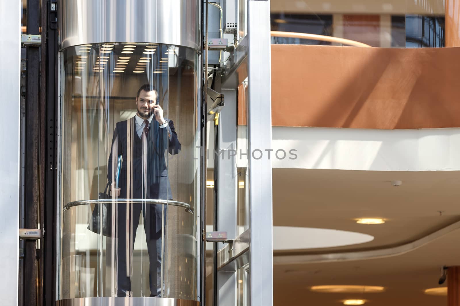 Businessman in modern glass elevator talking by the phone and holding briefcase