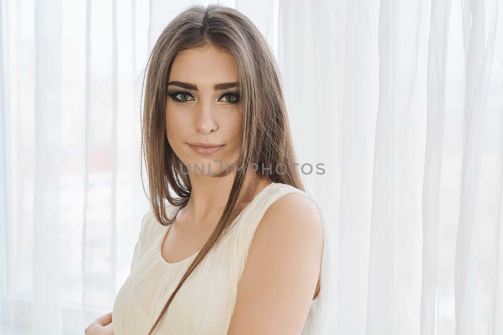 Cheerful smiling young white skin female with long brunette hair posing against the window in the morning, meeting a new day, dressed in a light transparent white tunic. Morning makeup.