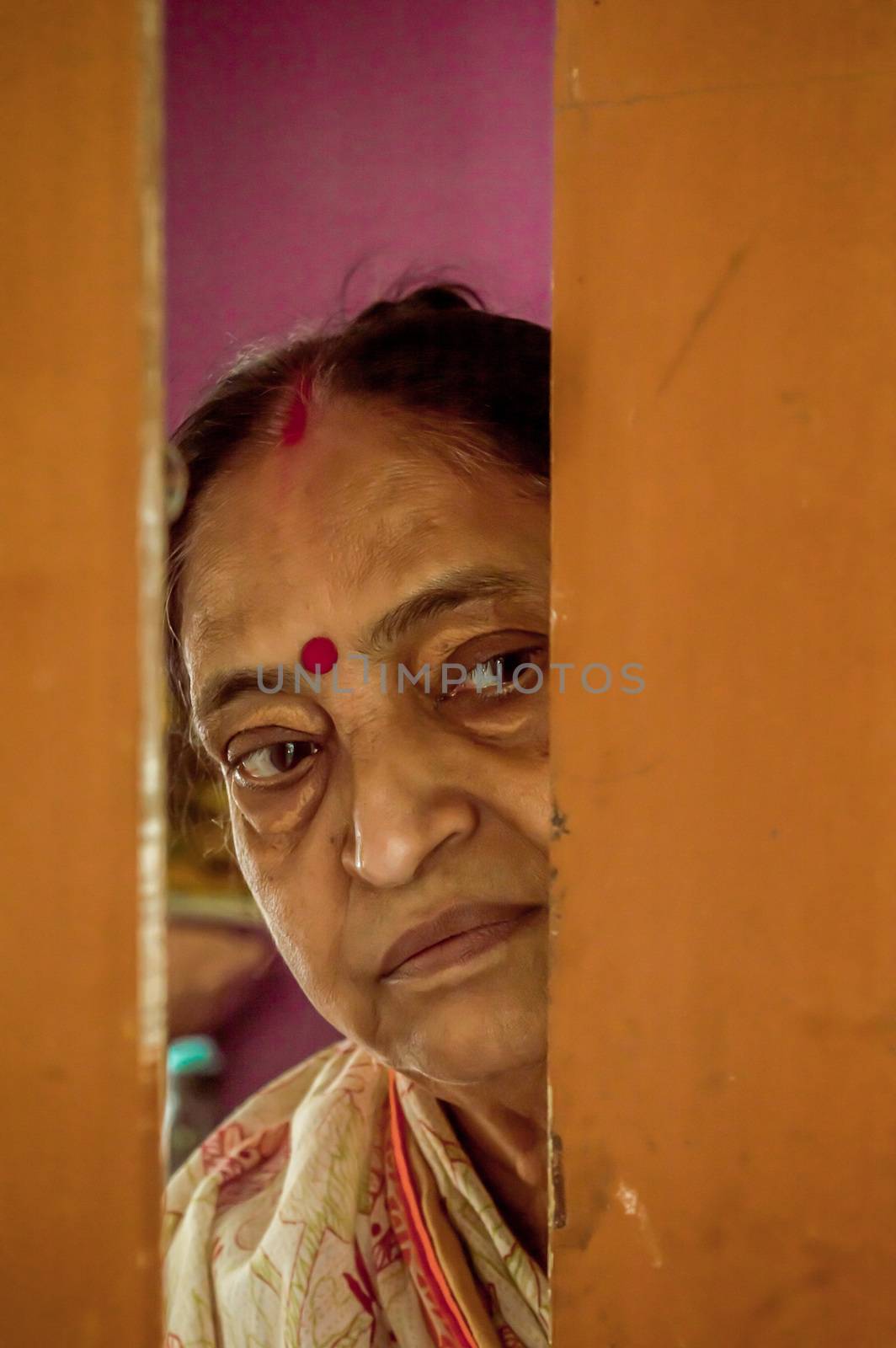 Thoughtful, sad, Worried and alone elderly senior woman peeping through the door at home and eagerly waiting for her son to come. Loneliness concept by sudiptabhowmick
