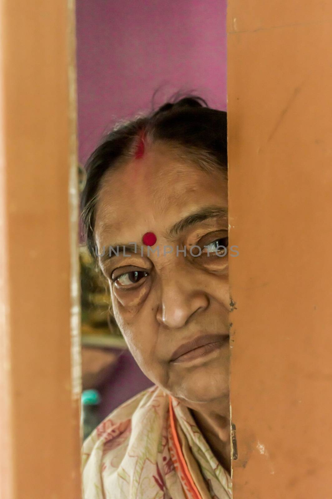 Thoughtful, sad, Worried and alone elderly senior woman peeping through the door at home and eagerly waiting for her son to come. Loneliness concept by sudiptabhowmick