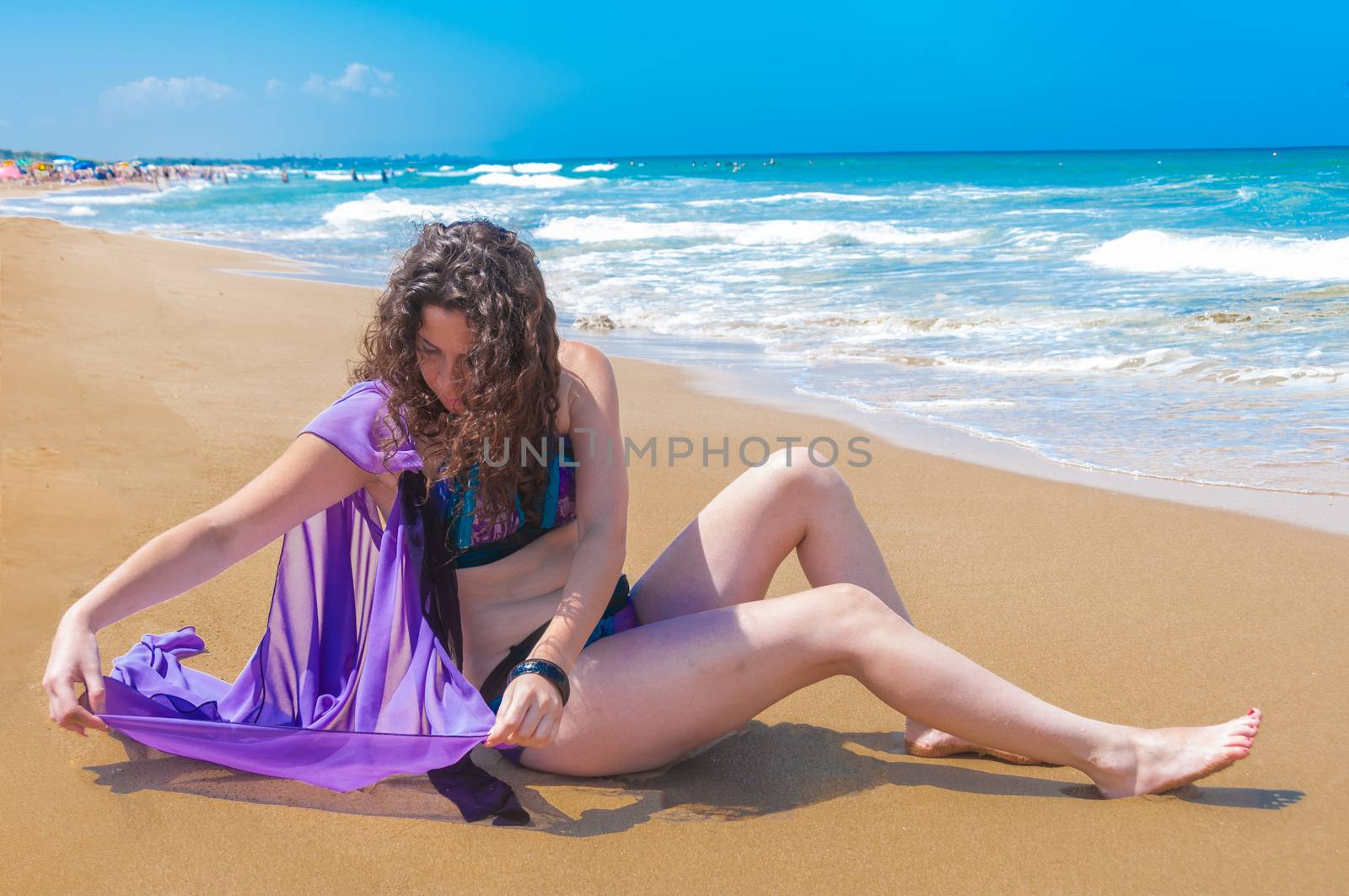 A girl sitting on a sandy beach, gently spreads a soaked purple, translucent cape.