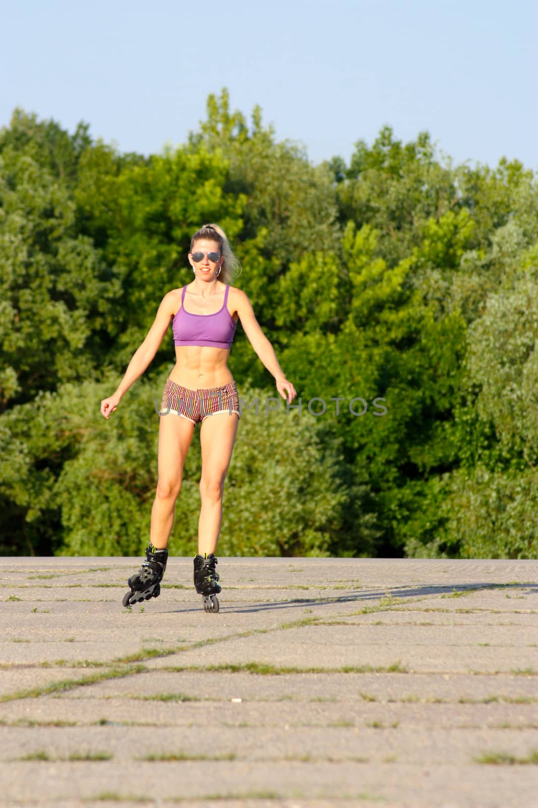 Blonde girl in sunglasses on roller-skates in sunny weather on an asphalt path on the green trees background