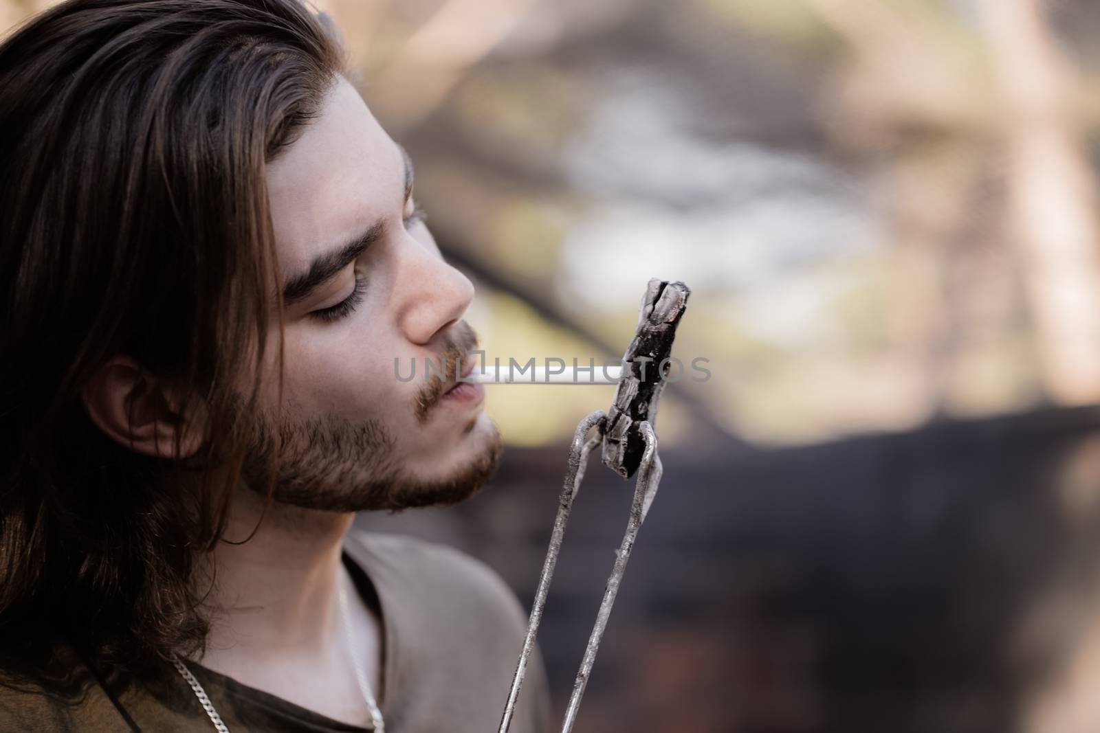 Portrait of a handsome young man with a beard, lighting a cigarette from charcoal, holding his by barbecue tongs.