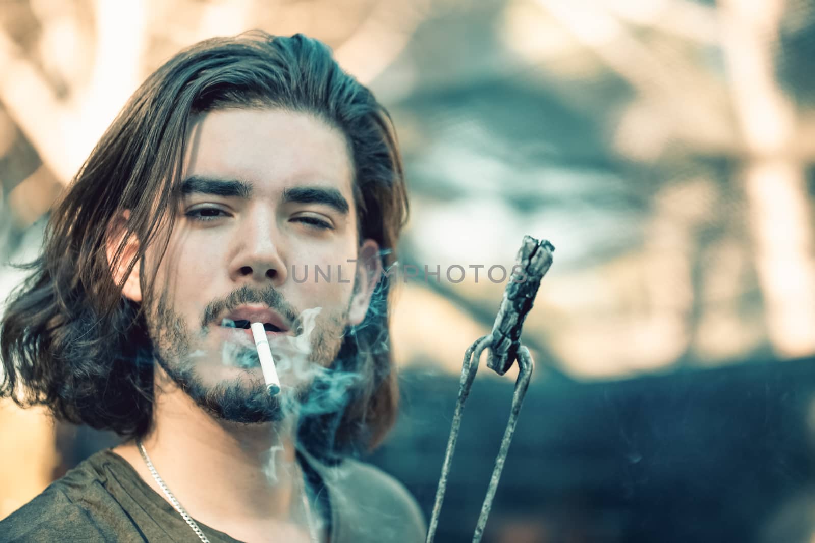 Portrait of a handsome young man with a beard, lighting a cigarette from charcoal, holding his by barbecue tongs.