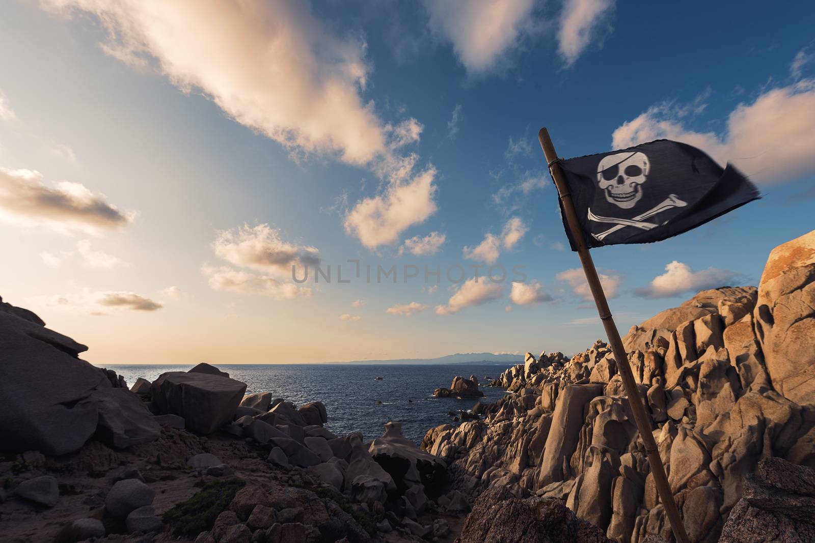 jolly roger flag flies at sunset pinned between the rocks of the pirate island in front of a few clouds in the crystal clear blue and orange sky, Capo Testa, Santa Teresa di Gallura, Sardinia, Italy