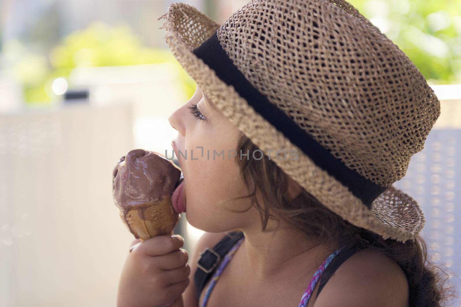 Little girl with a straw hat and a summer dress enjoys the summer heat eating a refreshing cone of chocolate ice cream, it melts in her hand while she licks it with her tongue