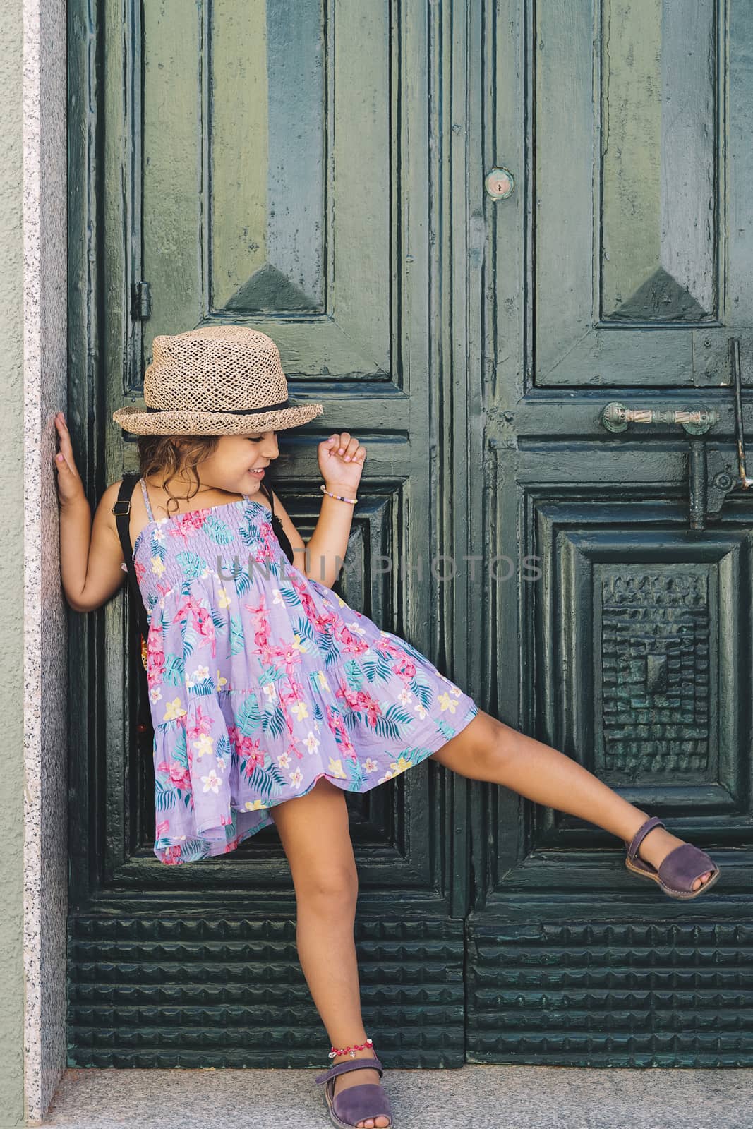 Little girl poses raising one leg in front of the green door of the house, she wears a lilac summer dress of flowers and a straw hat, vertical image
