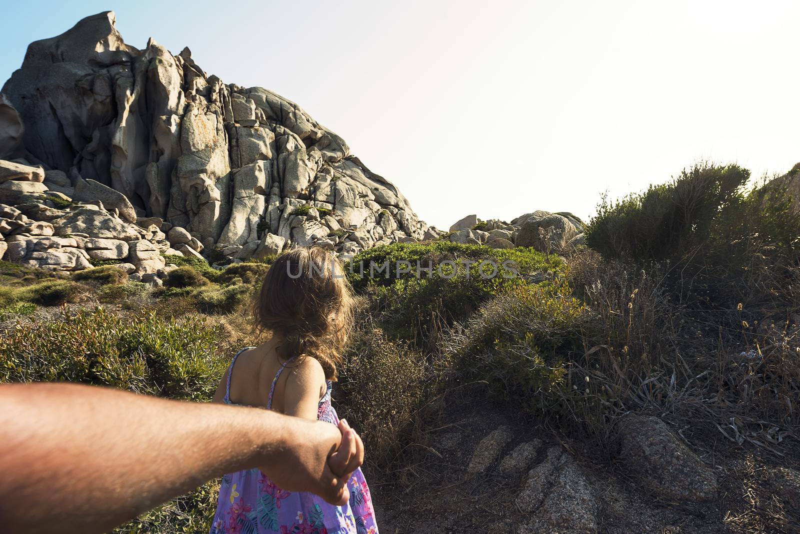 Kid strolling through the bushes holding her father's hand, in the background there are rocky mountains and the sky shines clear, subjective shot, Capo Testa, Santa Teresa di Gallura, Sardinia, Italy