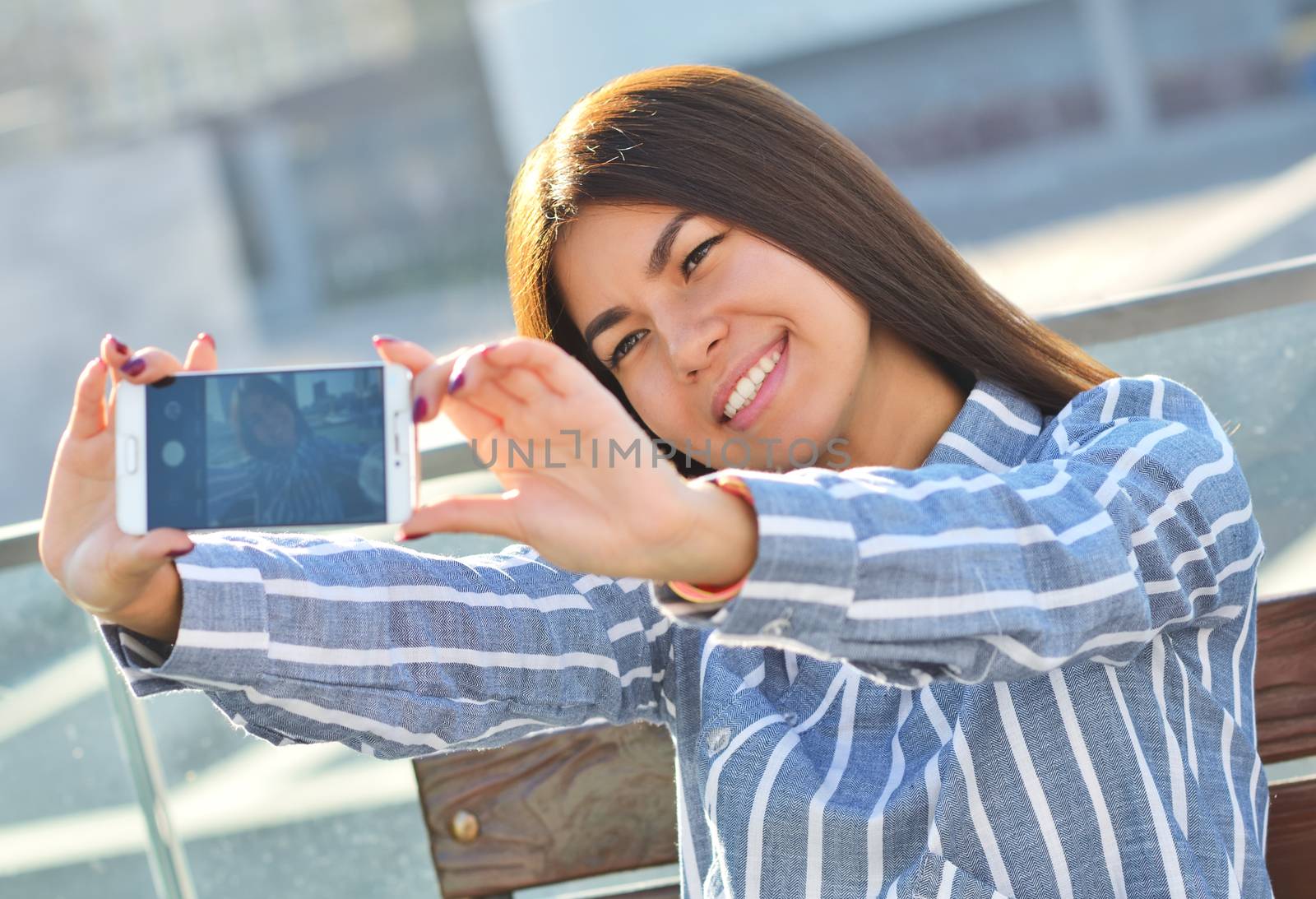 Young beautiful girl makes selfie and she is sitting on a bench in the city, the sunny day