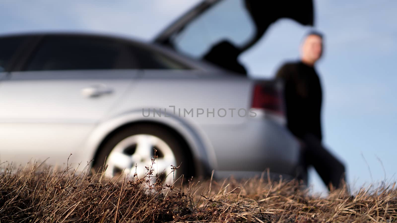 Sde view of young businessman in eyeglasses sitting on car trunk at nature - blurred background for banner