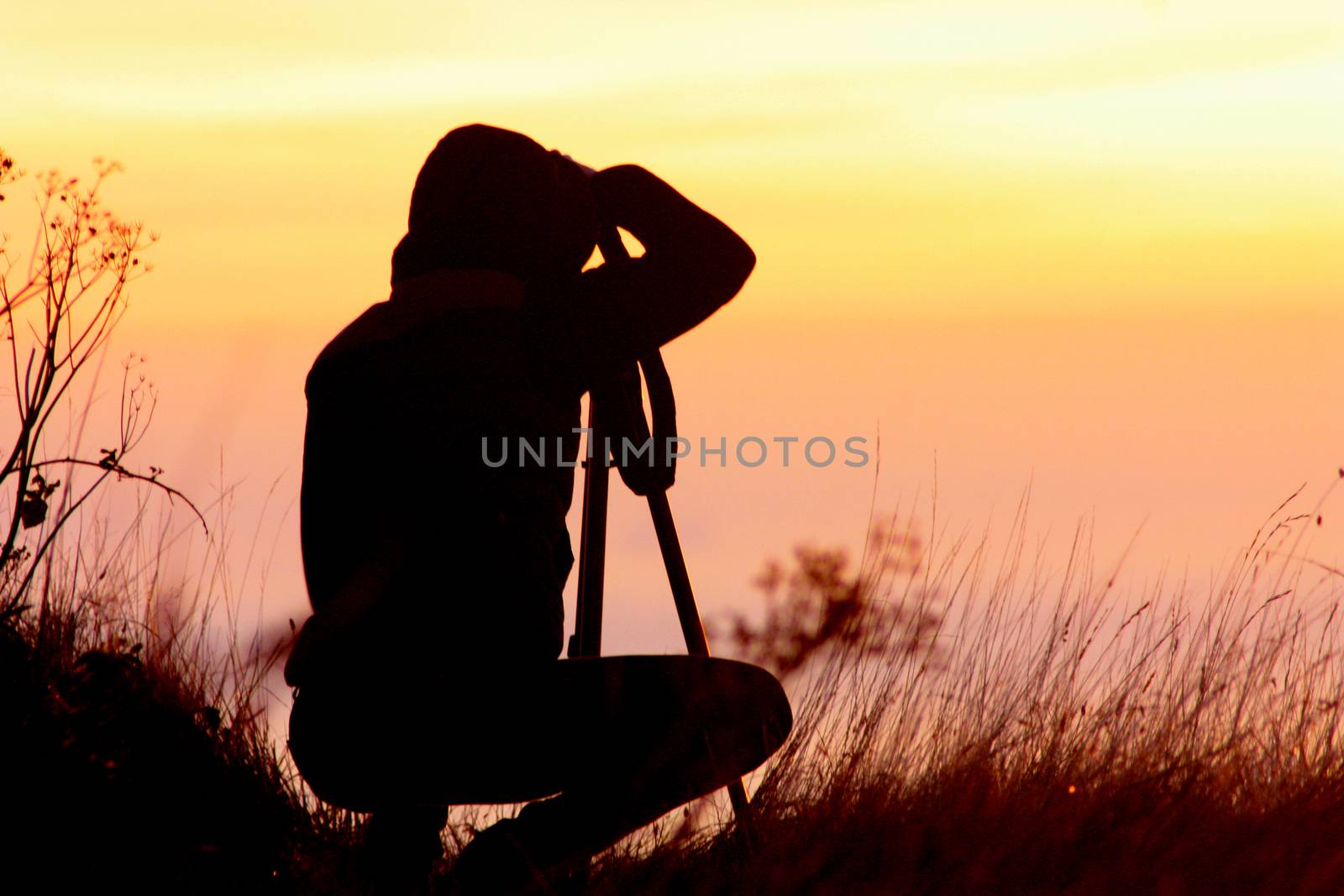 Silhouette of a girl photographer who is engaged in photography from a tripod against the background of an orange sunset.