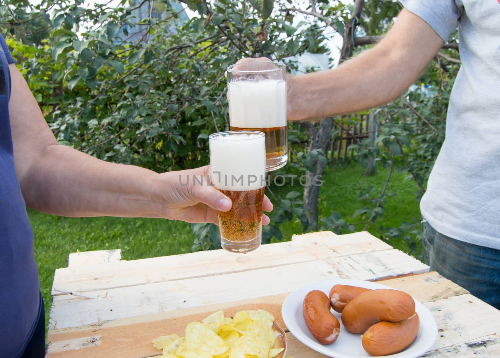 hands holding glasses with beer, chips and sausages lying on a light wooden boards, people relax in the weekends