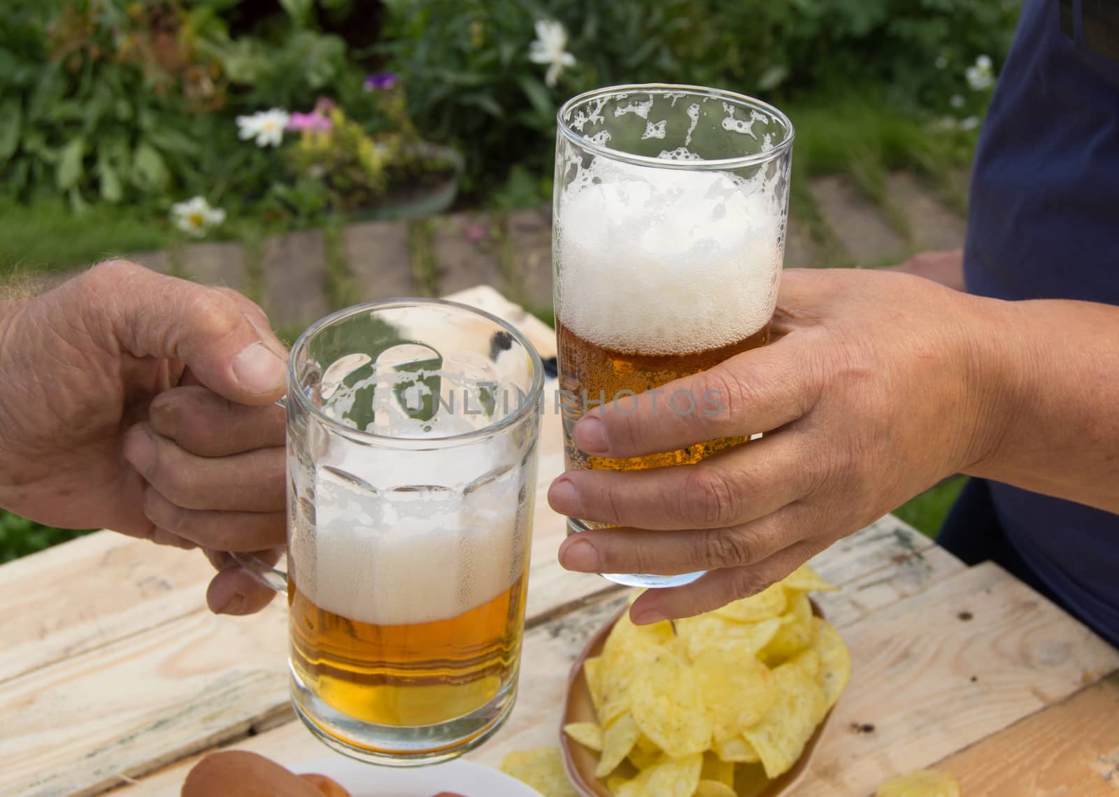 hands holding glasses with beer, chips and sausages lying on a light wooden boards, people relax in the weekends